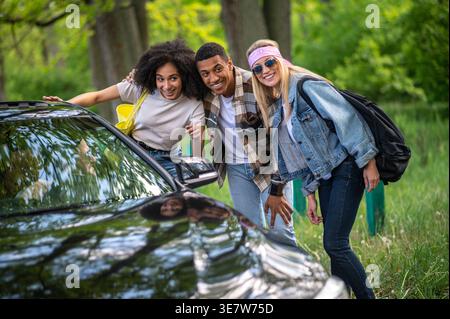 Smiling Black woman with male and female friends talking to the driver of black car pointing direction while standing on forest road on sunny day Stock Photo