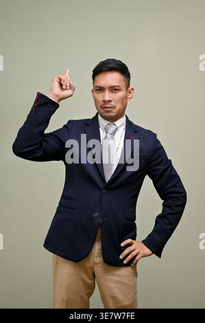 Serious Asian businessman pointing finger up standing with hand on hip against gray background Stock Photo