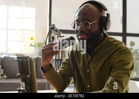 Black male podcaster holding and talking on microphone doing live stream while sitting at table in home studio during daytime Stock Photo