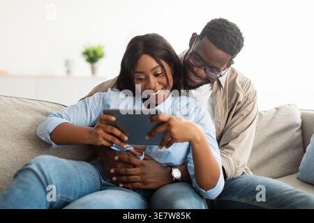 Black woman using tablet sitting with husband embracing her on sofa in living room at home Stock Photo