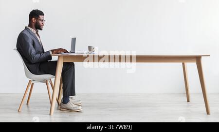 Black businessman wearing eyeglasses using laptop sitting at table with cup of coffee in front of white wall in office Stock Photo