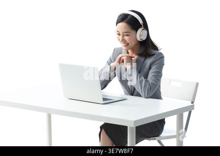 Smiling Chinese female teacher in headphones having video call using laptop while sitting at table against white background in studio Stock Photo