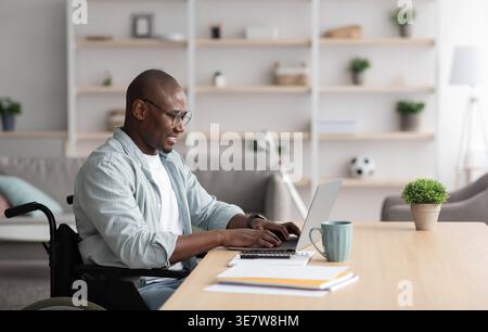 Smiling disabled Black bald businessman in wheelchair wearing eyeglasses working on laptop at table with smartphone and documents in living room at... Stock Photo