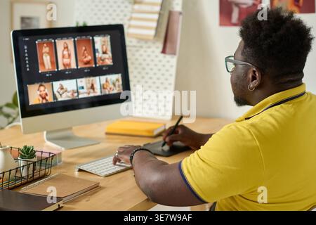 Black male photo editor wearing yellow polo shirt using graphic tablet for retouching photos on computer while sitting at table in office Stock Photo