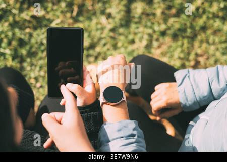 Hispanic woman wearing smartwatch sitting on bench with Hispanic female using smartphone in park on sunny day Stock Photo
