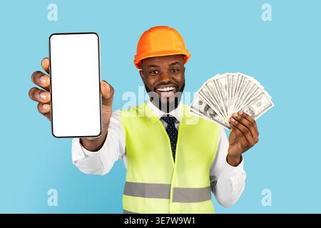 Black male engineer wearing hard hat and safety vest showing money and smartphone with blank screen while standing against blue background in studio Stock Photo