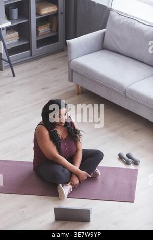 High-angle view of curvy Black woman working out sitting with crossed legs on yoga mat near laptop and dumbbells at home Stock Photo