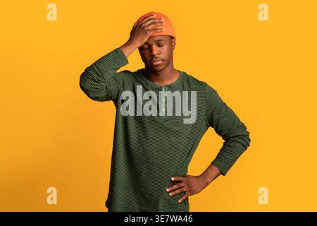 Black man in beanie touching head while standing with closed eyes and hand on waist against yellow background in studio Stock Photo