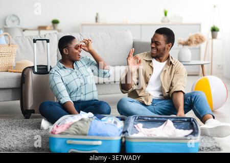 Cheerful Black man gesturing approving wifeâ€™s sunglasses while sitting on floor with packed suitcase in living room at home Stock Photo