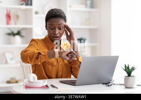 Shocked Black woman with short hair looking at her watch and touching head while sitting at table with laptop in office Stock Photo