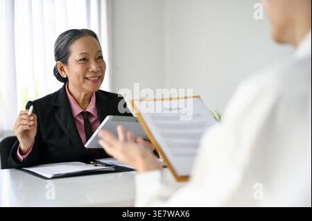 Asian male candidate sitting at table holding his resume and telling about his career experiences during job interview with senior Asian female HR ... Stock Photo