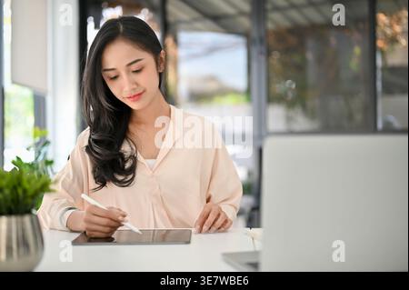 Smiling Asian business woman using stylus pen on tablet while sitting at table in office during daytime Stock Photo