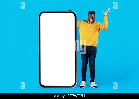 Excited Young Black Man Standing Near Huge Black Smartphone With White Screen, Surprised African American Guy Showing Big Empty Cellphone With Copy Sp Stock Photo