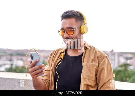 Hispanic man with beard wearing sunglasses and headphones using smartphone while standing outdoors during daytime Stock Photo