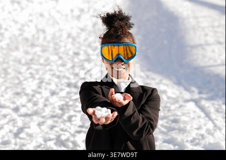 Smiling African American woman in goggles holding snowballs on both hands while standing on snowy mountain during winter Stock Photo