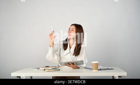 Smiling Asian woman pointing stylus pen up while holding tablet sitting at table with notebook and coffee cup in office Stock Photo