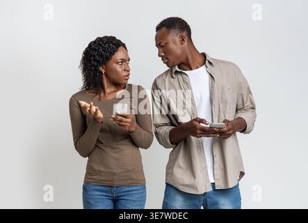 Black man holding smartphone spying on his girlfriend standing beside against grey background in studio Stock Photo