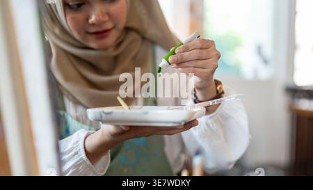Asian Muslim woman wearing hijab putting color on palette standing in art studio Stock Photo
