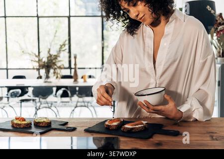 Hispanic woman preparing smoked salmon toast for breakfast standing in kitchen Stock Photo