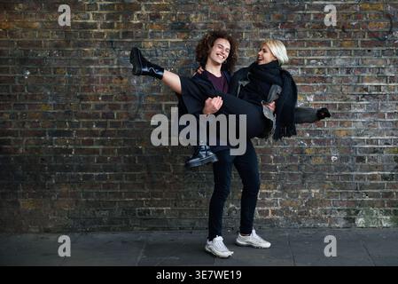 Cheerful Mixed race man with curly hair carrying caucasian girlfriend while standing against brick wall on street Stock Photo