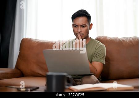 Confused and doubtful Asian man looking at his laptop with hand on mouth while sitting on sofa at home Stock Photo