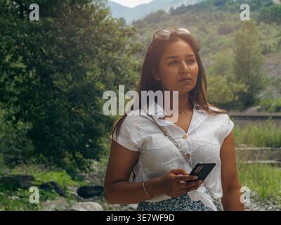 Serious Mixed race woman with eyeglasses on head holding smartphone looking away standing on rocks near river on sunny day Stock Photo