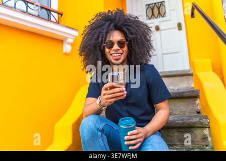 Hispanic man with afro wearing blue shirt and sunglasses holding water tumbler and using smartphone while sitting on stairs outside a yellow reside... Stock Photo