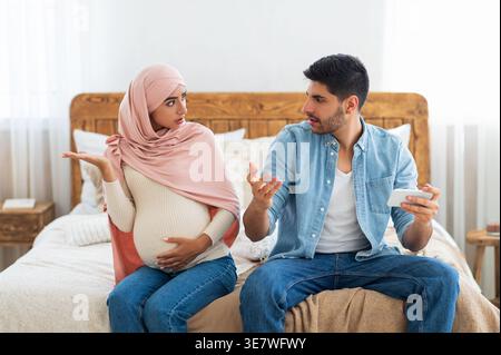 Pregnant Middle Eastern wife and Middle Eastern husband arguing while sitting on bed in bedroom at home Stock Photo