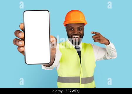 Black male engineer wearing hard hat and safety vest showing and pointing at smartphone with blank screen while standing against blue background in... Stock Photo