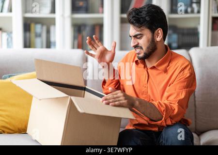 Disappointed Middle Eastern man gesturing hand opening parcel sitting on couch at home Stock Photo