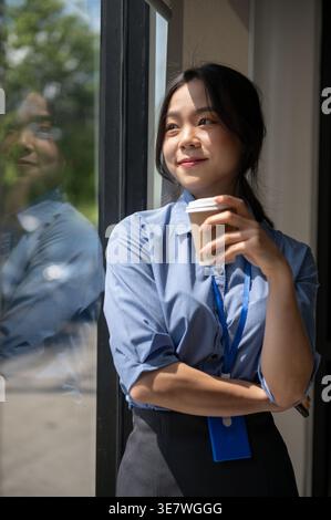 Smiling Asian female office worker having coffee looking out the window standing in office Stock Photo