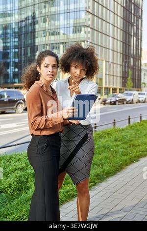 Mixed-race business woman pointing away showing something to female colleague holding tablet while standing together on city street on sunny day Stock Photo