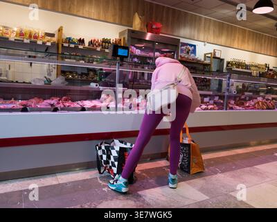 Woman in pink hoodie and purple leggings looking at fresh meat in a butcher shop display counter during shopping Stock Photo