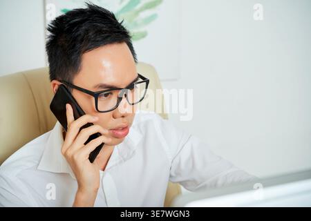 Asian male graphic designer having phone call using smartphone while sitting on chair in office Stock Photo