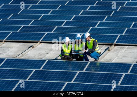 Diverse group of engineers wearing hard hats and safety vests discussing data on tablet while crouching and working on large solar panel installati... Stock Photo