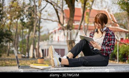 Asian female college student wearing eyeglasses and headphones around neck using tablet studying while sitting on bench in park during daytime Stock Photo