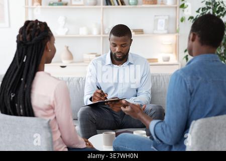 Black male psychologist having therapy session with couple and taking notes while sitting on couch in office Stock Photo