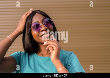 Smiling Black female in sunglasses touching hair and face looking away while standing against brown wall Stock Photo