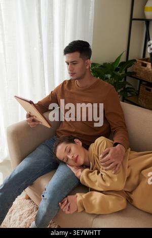 Mixed race woman sleeping and lying on lap of Asian boyfriend using tablet sitting on sofa in living room at home Stock Photo