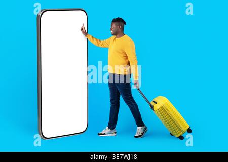 Black man with luggage using big smartphone while standing in front of blue background in studio Stock Photo