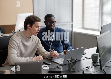 Black and white male IT specialists working on laptop together sitting at desk in office Stock Photo