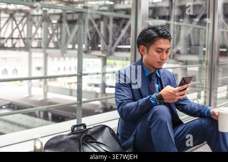Asian businessman in blue suit using smartphone while sitting on floor with coffee at train station during daytime Stock Photo
