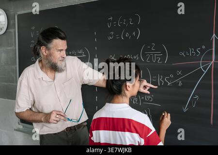 Asian female student solving math problem on blackboard with middle-aged caucasian male teacher helping her while standing in the classroom Stock Photo