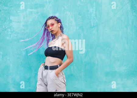 Black woman with purple braided hair standing with hands in pockets against turquoise wall Stock Photo