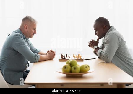 Black man and middle-aged caucasian male friend playing chess while sitting at table with green apples at home Stock Photo