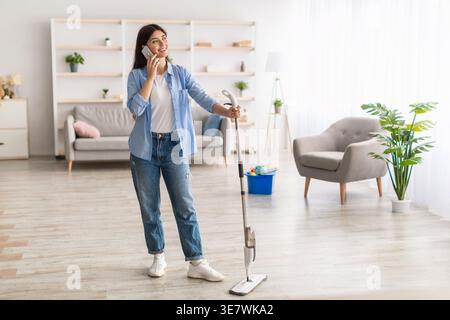 Middle Eastern woman having phone call looking away holding mop while standing in living room at home Stock Photo