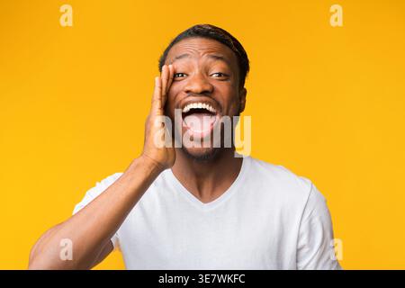 Hey. Positive African American Man Shouting Holding Hand Near Mouth Standing On Yellow Studio Background Stock Photo