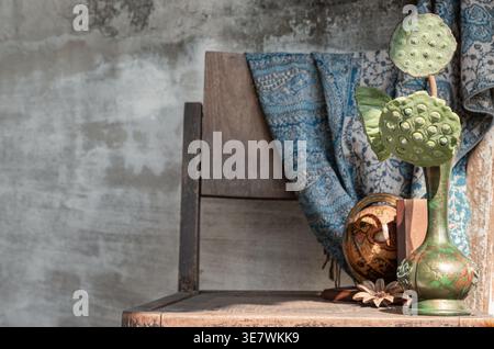 On a weathered wooden chair, an ornate green-tinted bronze vase holds two dried lotus seed pods, anchored against a backdrop of a textured concrete wa Stock Photo