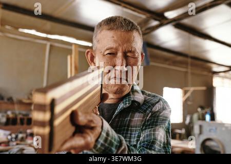 Senior Asian male carpenter squinting eyes when checking evenness of wooden plank while standing in workshop Stock Photo
