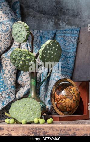 A vintage copper vase holds a cluster of green lotus seed pods, their long stems curving elegantly against a patterned fabric backdrop. To the right s Stock Photo